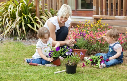 A woman with short blonde hair and wearing a white blouse is kneeling on a neatly maintained grassy lawn in a garden, engaged in planting and tending to a variety of potted flowers, including purple and pink blooms. She is accompanied by two young children, both with light brown hair and dressed in casual clothing; one is sitting on the grass holding a potted plant, while the other is using a small gardening tool to dig in a separate pot. The garden features a lush border of large, variegated foliage plants behind them, alongside a wooden deck with a railing in the background. The scene is outdoors on a bright, clear day, with natural light illuminating the garden area, emphasizing the vibrant colours of the flower plants and the well-kept grass. The environment reflects a typical residential garden designed for family outdoor activities, with an emphasis on gardening and plant care, aligning with local landscaping services such as lawn and garden maintenance provided by Lawn Mowing Yeading in the area near Yeading, Middlesex, UB5 postcode.